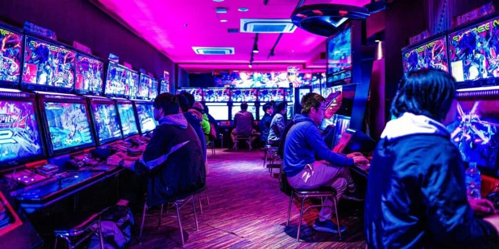 ผู้คนนั่งเล่นเกมอาร์เคดในห้องที่มีแสงไฟสีชมพูและม่วง